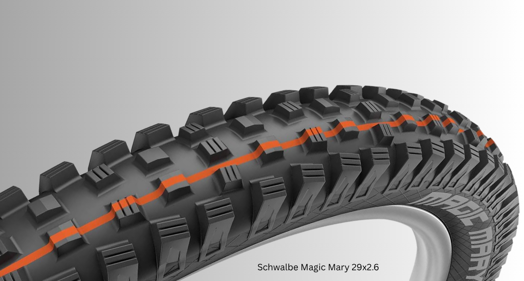 Close-up of a Schwalbe Magic Mary 29x2.6 mountain bike tire featuring aggressive black knobs and a distinctive orange Addix Soft compound stripe down the center.