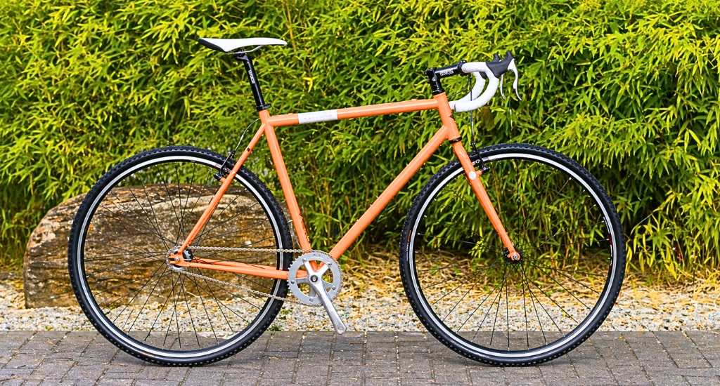 An orange and white Genesis Day One single-speed road bike with drop handlebars and knobby tires, parked on a stone path against a lush green bamboo background.
