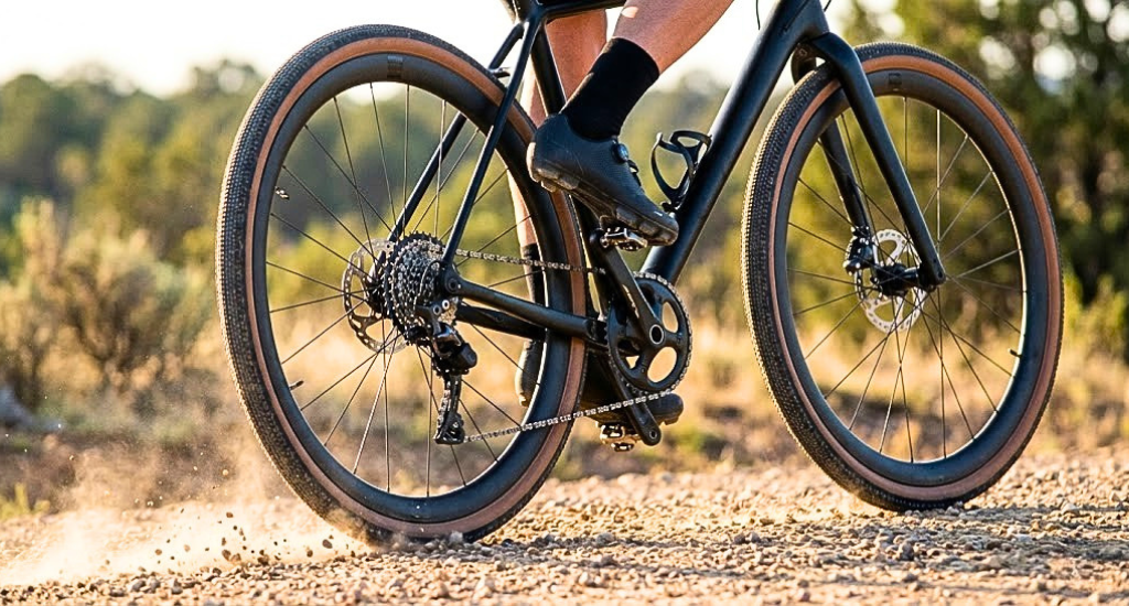 A cyclist rides a sleek black gravel bike on a dusty trail. The focus is on the rear wheel kicking up dirt, highlighting the tan-wall tires and disc brakes.