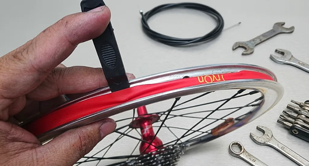 Hand using a black tire lever to align red rim tape inside an aluminum bicycle wheel channel.
