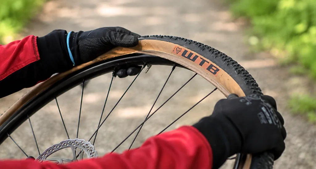 Gloved hands mounting and stretching a WTB tan sidewall gravel tire bead onto a black bike rim outdoors.