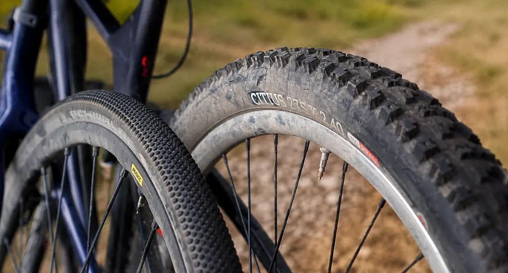 Close-up view of two bike wheels showing narrow gravel tire tread alongside a wide knobby mountain bike tire mounted on a rim.